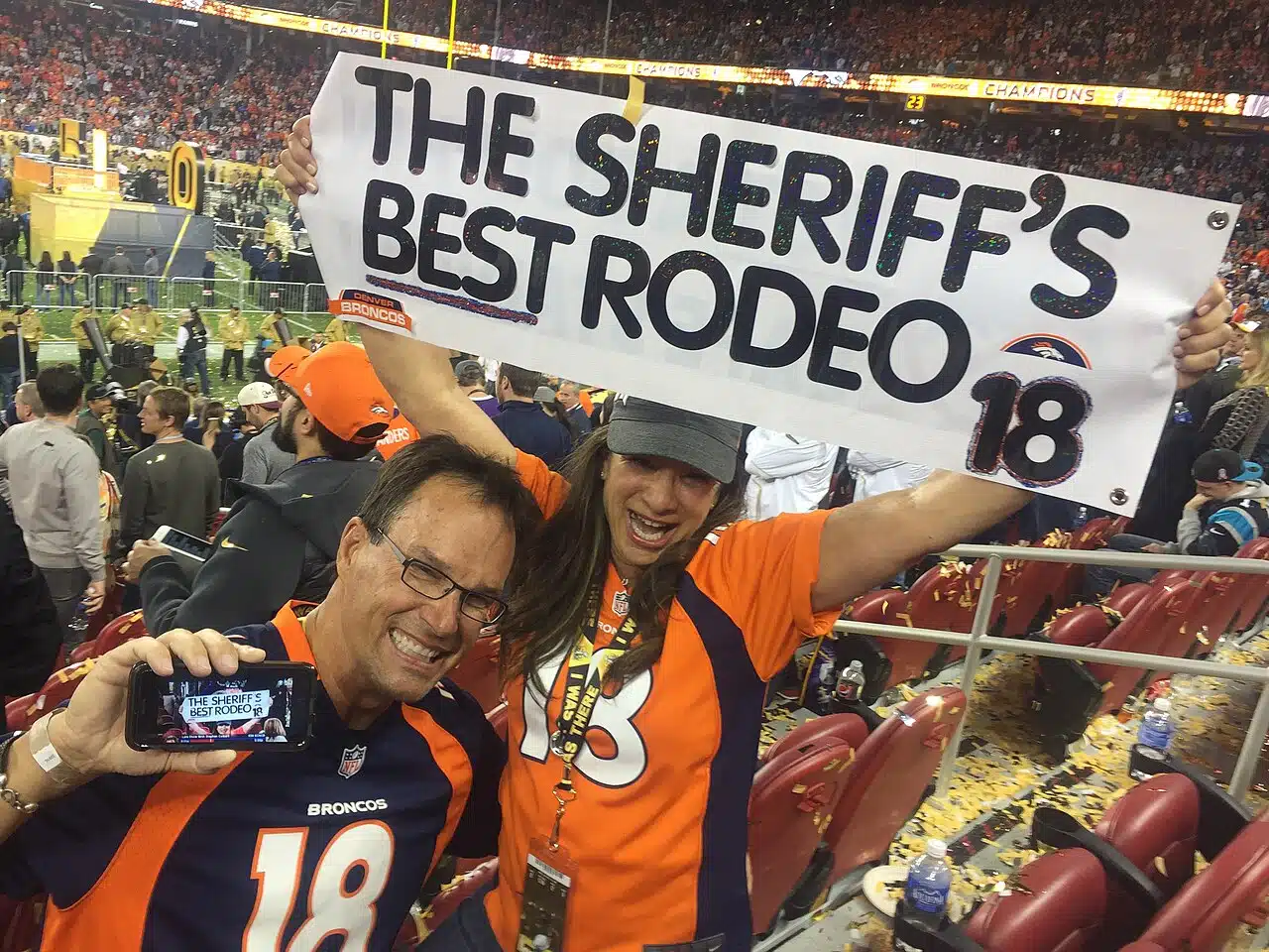Broncos fans in orange jerseys cheer and hold up a homemade sign in a crowd at Super Bowl 50.